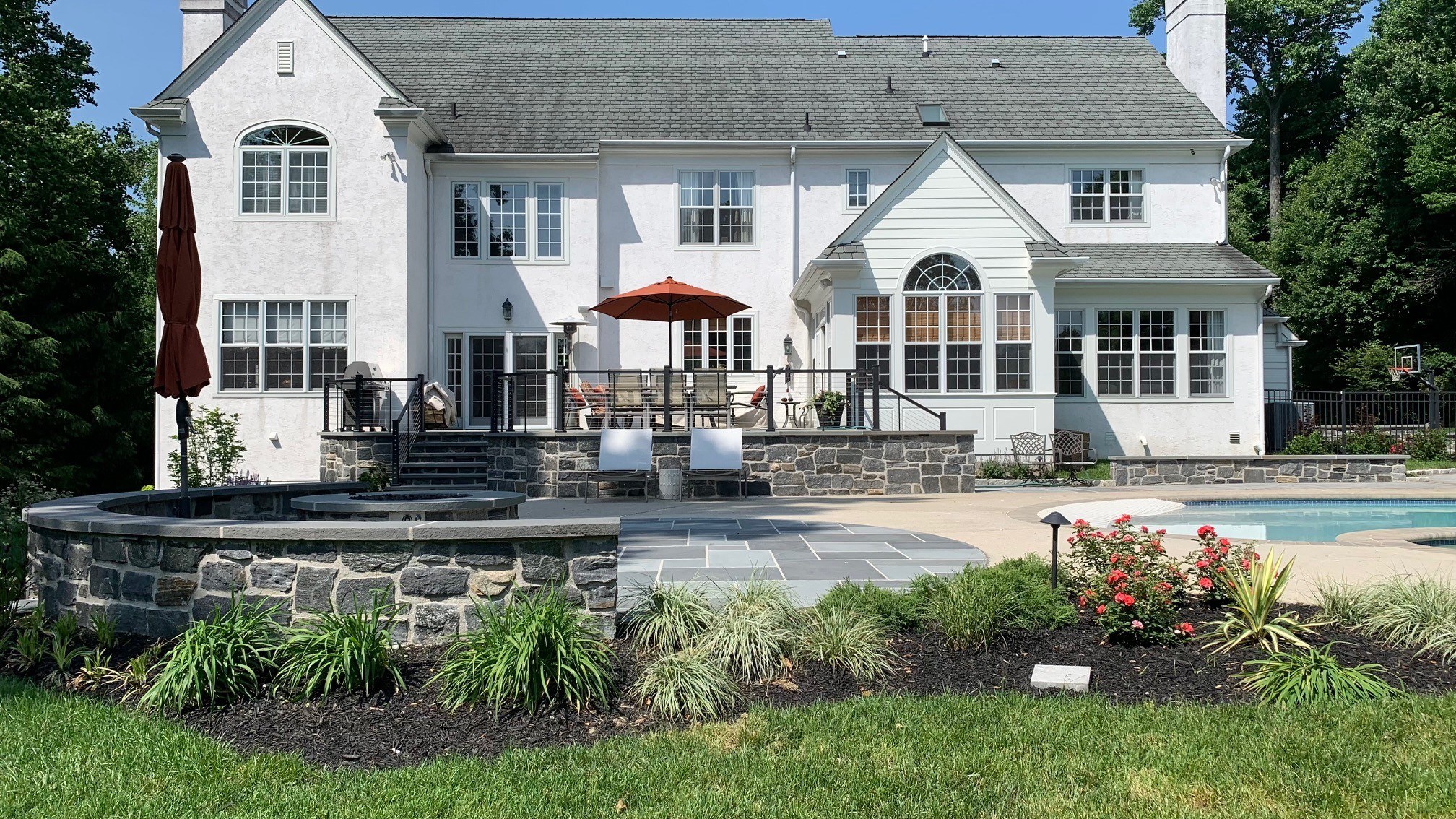 Backyard of a white Main Line Colonial home with stone-walled patio, bluestone hardscape, perennial gardens and an inground pool by JHL Pools in Chester County, PA.